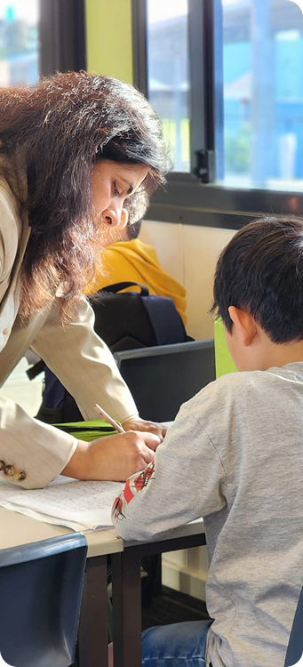Teacher with long hair leaning over a student in gray shirt sitting at a desk in classroom with window in background