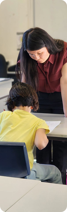 Teacher in maroon shirt leaning over a student in yellow shirt sitting at a desk in classroom